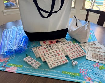 May include: A white tote bag with black handles and a black bottom sits next to a white pouch. Several sets of Mahjong tiles are arranged on a floral-patterned mat. Dice and instruction cards are also visible.