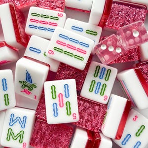May include: A collection of white Mahjong tiles with red, blue, and green markings. The tiles are scattered among red, textured tiles and two pink dice. The game pieces are for the classic tile-based game.