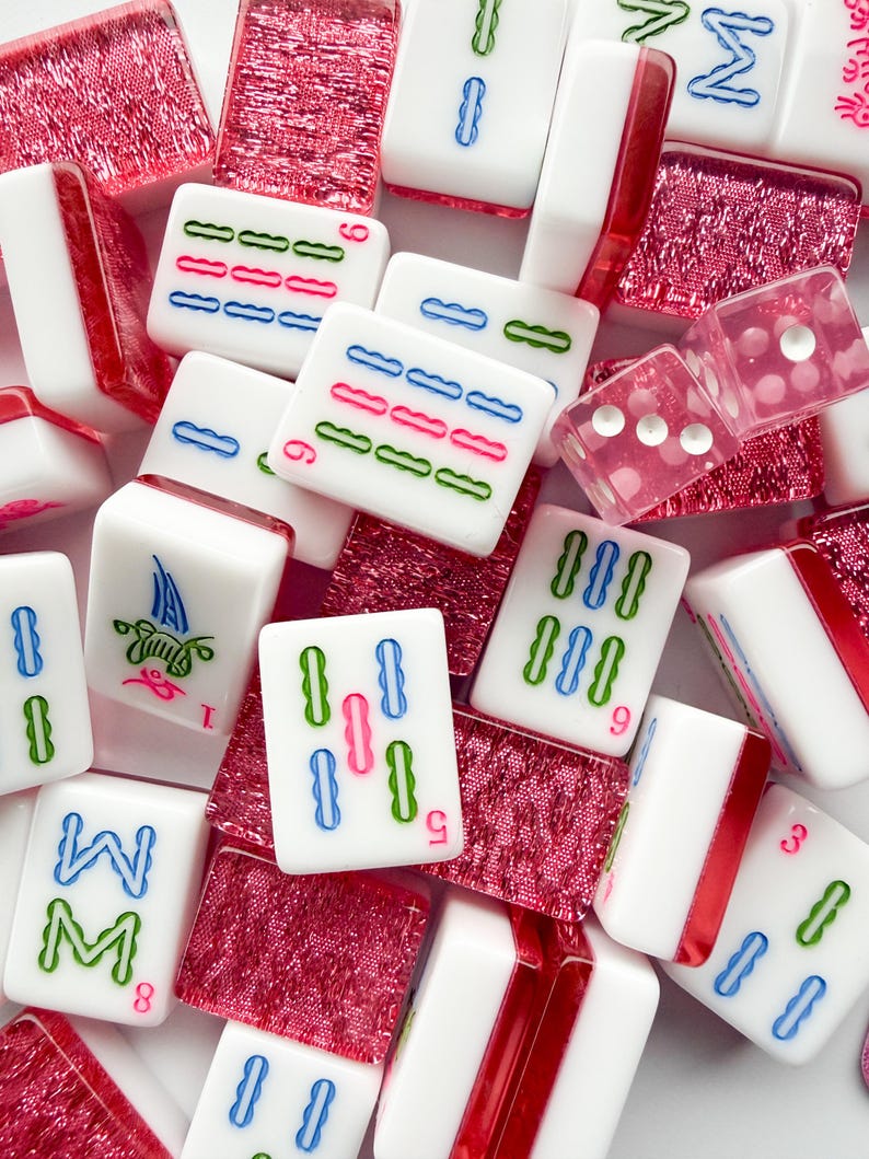 May include: A collection of white Mahjong tiles with red, blue, and green markings. The tiles are scattered among red, textured tiles and two pink dice. The game pieces are for the classic tile-based game.