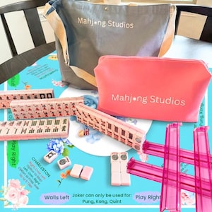 May include: A Mahjong set with pink tiles, dice, and accessories, including a gray tote bag and a pink zippered pouch, both with "Mahjong Studios" printed on them. The set is arranged on a blue floral patterned surface.