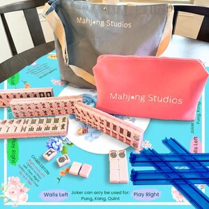 May include: A Mahjong game set with a gray tote bag and a pink zippered pouch, both with "Mahjong Studios" printed on them. The set includes pink tiles, dice, and blue tile racks, all arranged on a blue floral patterned surface.