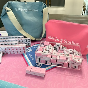May include: A collection of Mahjong Studios game sets. The image features a light blue tote bag and a pink zippered pouch, both with the text "Mahjong Studios." The set includes pink and white tiles, some in a clear acrylic container, and others in clear plastic cases.