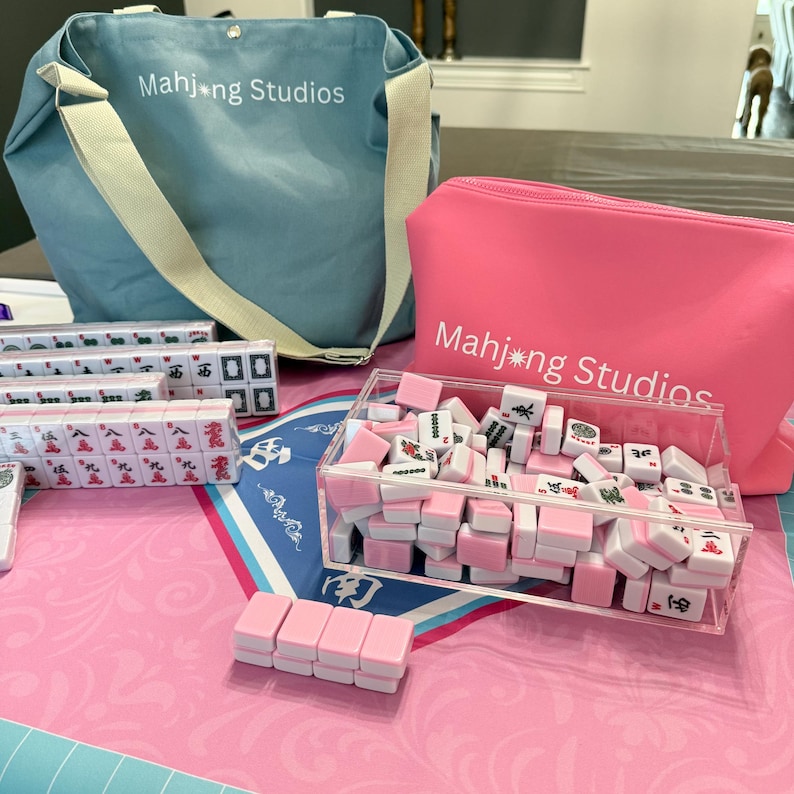 May include: A collection of Mahjong Studios game sets. The image features a light blue tote bag and a pink zippered pouch, both with the text "Mahjong Studios." The set includes pink and white tiles, some in a clear acrylic container, and others in clear plastic cases.