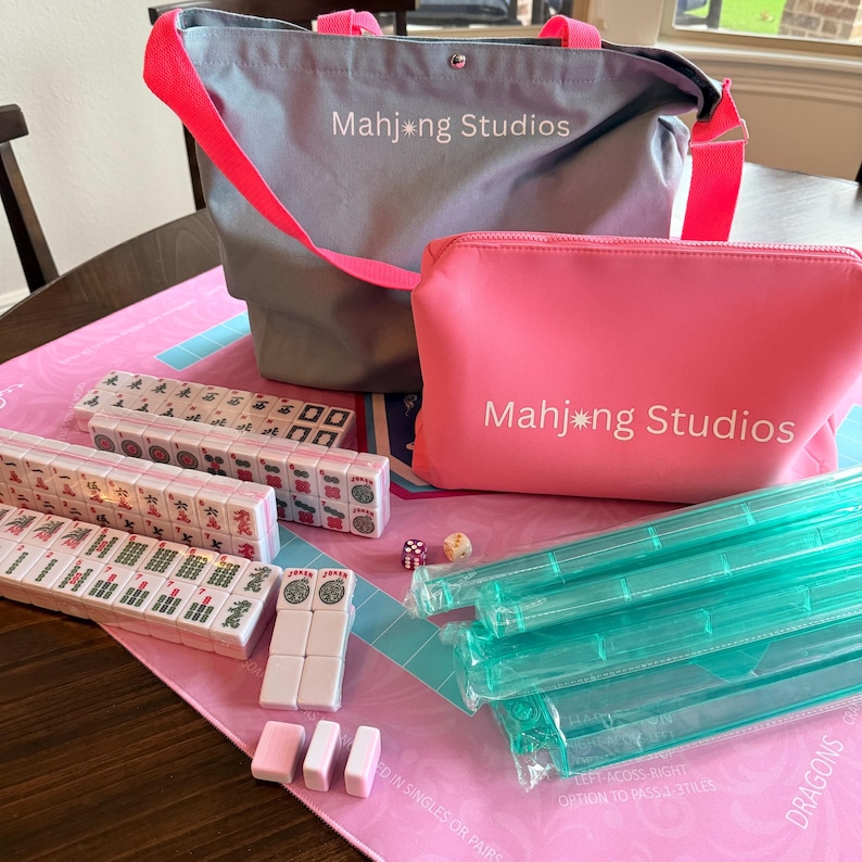 May include: A Mahjong set featuring a pink playing mat, tiles, dice, and teal plastic racks. The set includes a grey tote bag and a pink zipped pouch, both with "Mahjong Studios" printed on them. The set is ready for play.