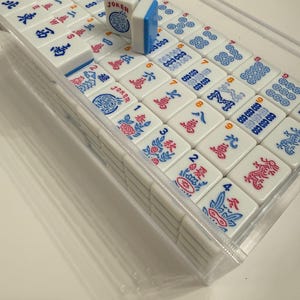 Puede incluir: Un juego de fichas de Mahjong blancas con s&iacute;mbolos y caracteres azules y rojos, presentado en una caja de pl&aacute;stico transparente. Una ficha est&aacute; de pie, mostrando la palabra "JOKER". Las fichas est&aacute;n dispuestas en filas y columnas.