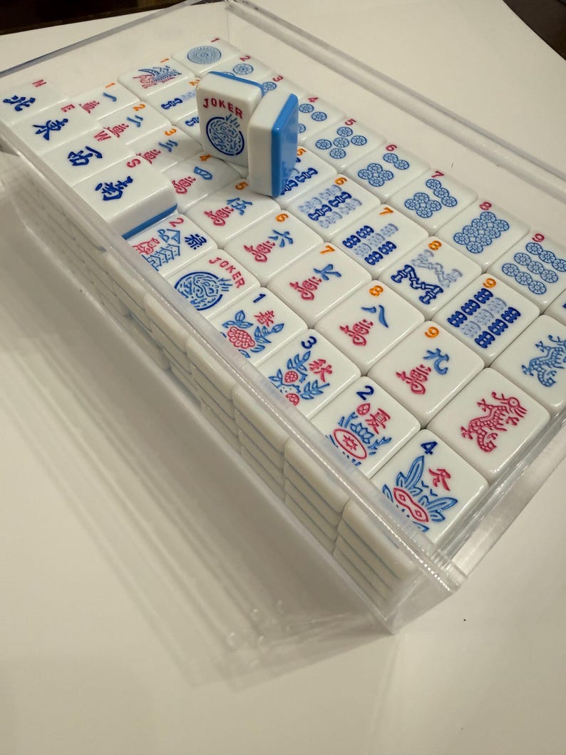 Puede incluir: Un juego de fichas de Mahjong blancas con s&iacute;mbolos y caracteres azules y rojos, presentado en una caja de pl&aacute;stico transparente. Una ficha est&aacute; de pie, mostrando la palabra "JOKER". Las fichas est&aacute;n dispuestas en filas y columnas.