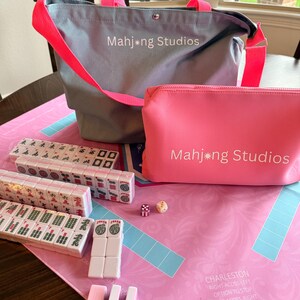 May include: A Mahjong set featuring a light blue tote bag and a pink cosmetic bag, both displaying "Mahjong Studios". The set includes tiles, dice, and a pink game mat with blue details.