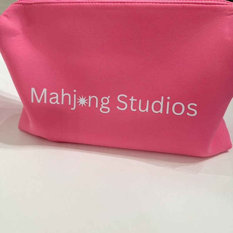 May include: A bright pink cosmetic bag with the text "Mahj*ng Studios" printed in white. The bag is made of a soft material and has a zipper closure. The bag is sitting on a white surface.