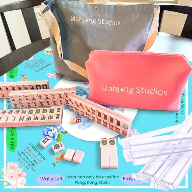 May include: A Mahjong set with pink and white tiles, dice, and accessories. The set includes a gray tote bag and a pink cosmetic bag, both with "Mahjong Studios" printed on them. The game is set on a blue floral patterned surface.