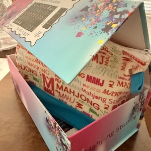 May include: A box with a pink and blue gradient exterior, containing a Mahjong Studios branded pouch and a blue headband. The pouch is beige with red and white "MAHJONG" text. The box lid has a "Neon Shadow" label.