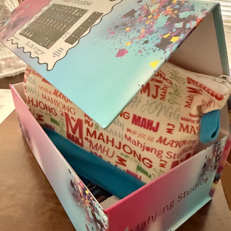 May include: A box with a pink and blue gradient exterior, containing a Mahjong Studios branded pouch and a blue headband. The pouch is beige with red and white "MAHJONG" text. The box lid has a "Neon Shadow" label.