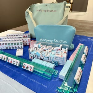 May include: A complete Mahjong set with tiles, racks, and accessories. The set includes a blue tote bag and a matching pouch, both with the text "Mahjong Studios". The tiles are white and blue, and the racks are clear and green.