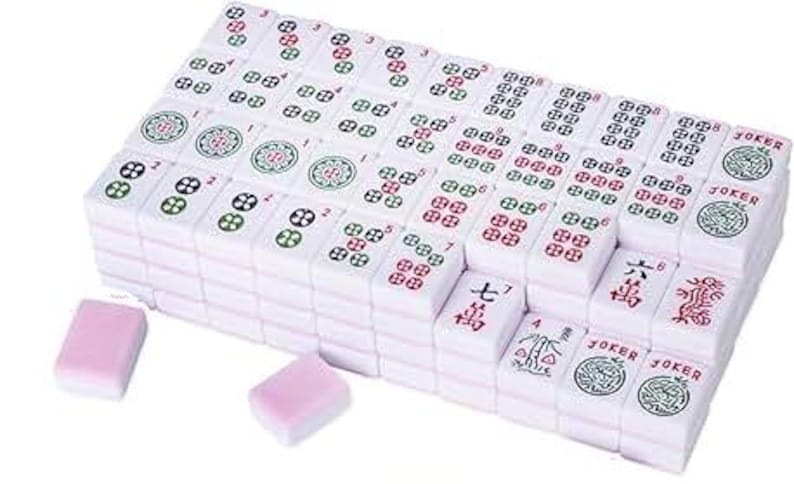 May include: A complete Mahjong set with white tiles featuring red, green, and black symbols and numbers. The set includes two pink dice. The tiles are stacked and arranged, ready for play.