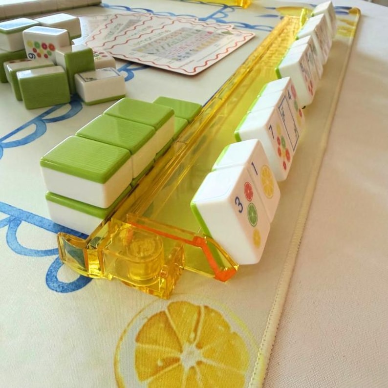 May include: A board game set featuring white and lime green rectangular tiles with citrus fruit illustrations and numbers. The tiles are arranged on a translucent yellow plastic rack, with additional tiles stacked nearby on a mat with lemon graphics.