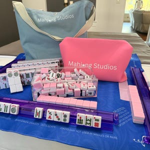 May include: A Mahjong set featuring pink and white tiles, a blue tote bag, and a pink pouch, all branded with the "Mahjong Studios" logo. The set includes a clear acrylic tile holder and a purple tile rack, set on a blue mat.