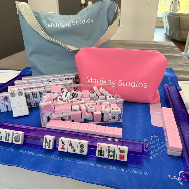 May include: A Mahjong set featuring pink and white tiles, a blue tote bag, and a pink pouch, all branded with the "Mahjong Studios" logo. The set includes a clear acrylic tile holder and a purple tile rack, set on a blue mat.