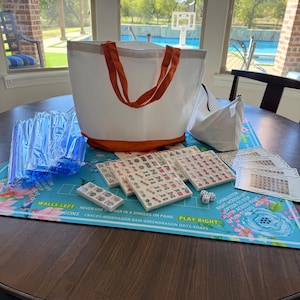 May include: A complete Mahjong set on a table. The set includes tiles, dice, and a blue floral play mat. A white tote bag with orange handles and base is also present, along with a smaller white bag. The game is ready to play.