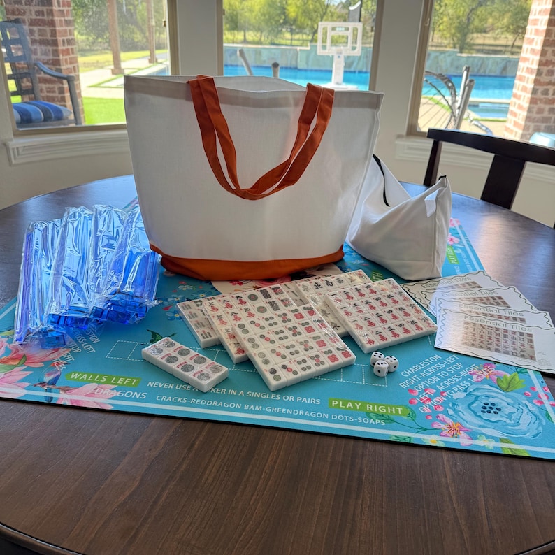 May include: A complete Mahjong set on a table. The set includes tiles, dice, and a blue floral play mat. A white tote bag with orange handles and base is also present, along with a smaller white bag. The game is ready to play.