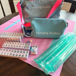 May include: A Mahjong set with tiles, dice, and accessories. The set includes a grey tote bag and a matching pouch, both with "Mahjong Studios" printed on them. The tiles are white with black and red markings. A pink mat and green plastic tile racks are also included.