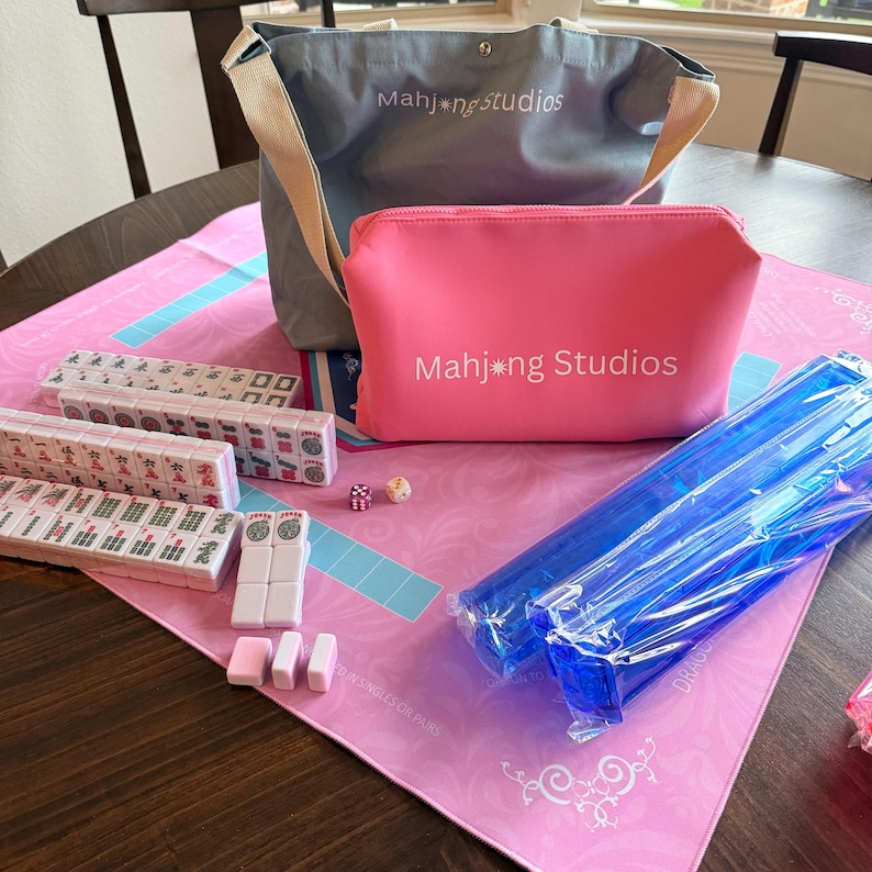 May include: A Mahjong set with a pink and blue theme. The set includes tiles, dice, a pink zippered pouch, a blue tote bag, and a pink playing mat. The text "Mahjong Studios" is visible on the bag and pouch.