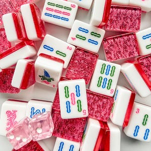 May include: A collection of white and red Mahjong tiles with various symbols and numbers in green, blue, and red. The set includes pink dice and red decorative pieces. The tiles are arranged in a scattered pattern.