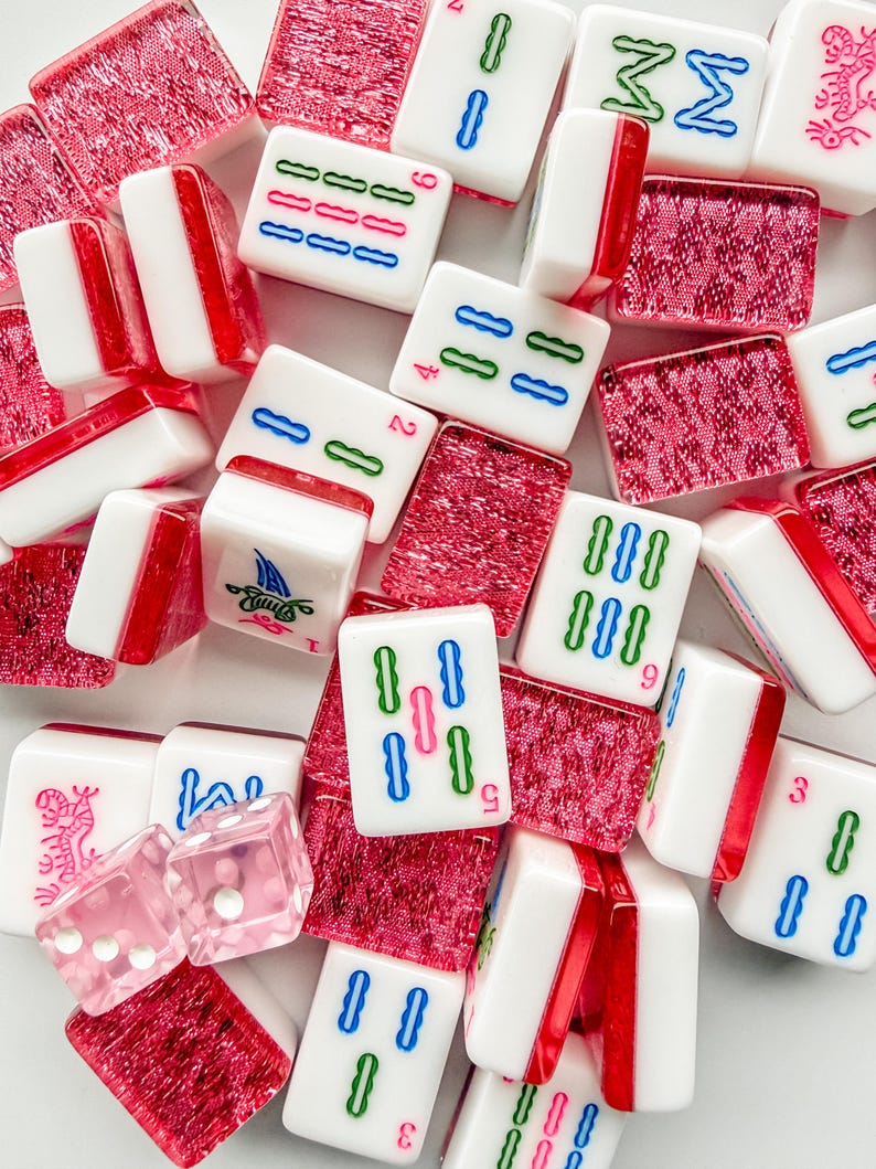 May include: A collection of white and red Mahjong tiles with various symbols and numbers in green, blue, and red. The set includes pink dice and red decorative pieces. The tiles are arranged in a scattered pattern.