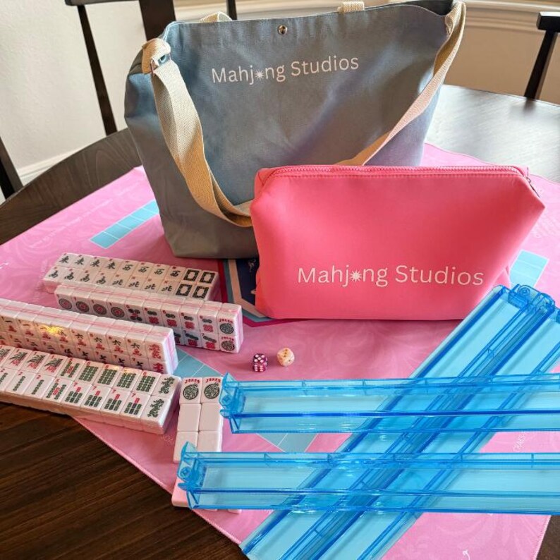 May include: A Mahjong set with a blue tote bag and a pink zippered pouch, both with "Mahjong Studios" printed on them. The set includes tiles, dice, and blue tile racks, all arranged on a pink mat.