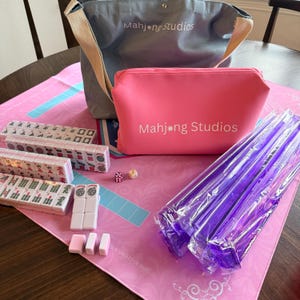 May include: A complete Mahjong set featuring tiles, dice, and accessories. The set includes a pink zippered pouch and a gray tote bag, both with "Mahjong Studios" printed on them. The tiles are arranged on a pink mat.
