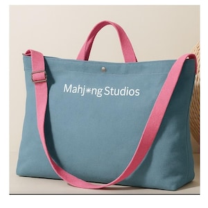 May include: A blue tote bag with pink straps and handles. The bag has the text "Mahj*ng Studios" printed on the front. The bag has a shoulder strap and a top handle. The bag is made of a canvas material.
