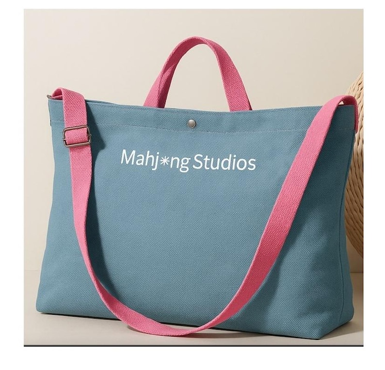May include: A blue tote bag with pink straps and handles. The bag has the text "Mahj*ng Studios" printed on the front. The bag has a shoulder strap and a top handle. The bag is made of a canvas material.