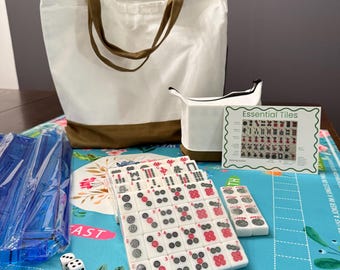 Könnte beinhalten: Ein komplettes Mahjong-Set mit weißen Spielsteinen mit roten und schwarzen Markierungen, Würfeln und einer Tragetasche. Die Tasche ist weiß mit braunen Griffen und Besatz. Ein kleines weißes Täschchen und eine Karte mit der Aufschrift "Essential Tiles" sind ebenfalls enthalten.