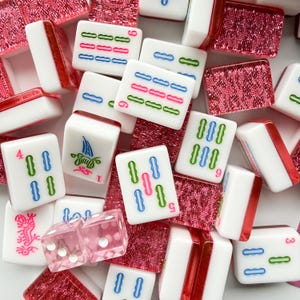 May include: A collection of white Mahjong tiles with colorful markings and red accents. The tiles feature blue, green, and pink symbols. Also included are pink dice and red decorative pieces.