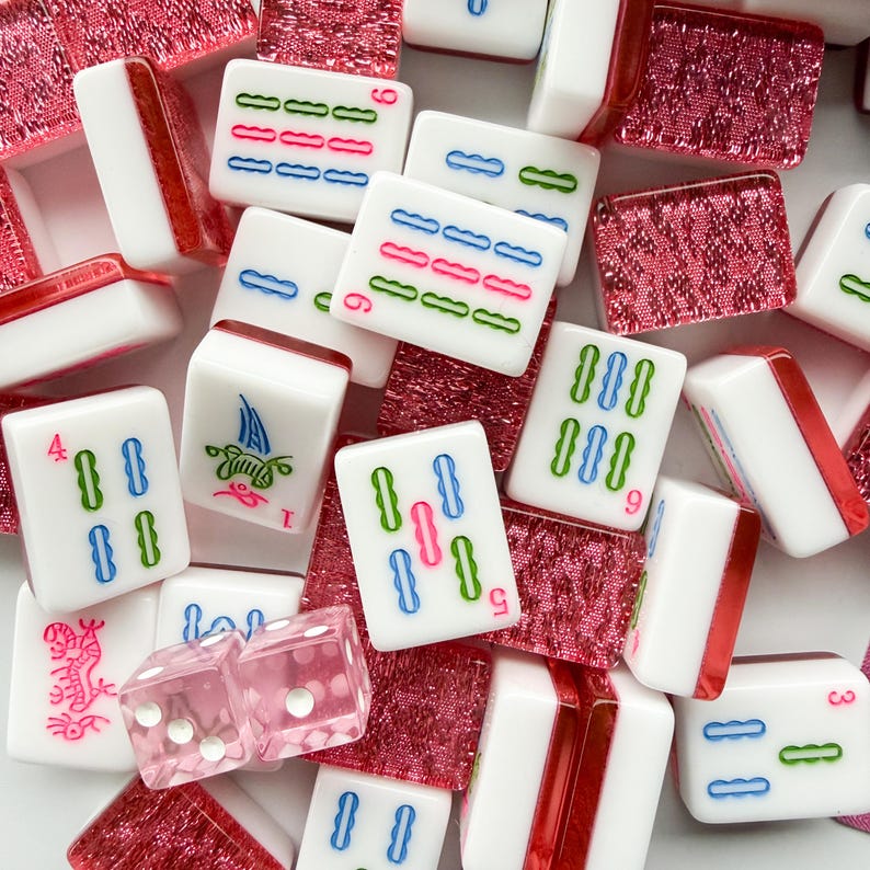 May include: A collection of white Mahjong tiles with colorful markings and red accents. The tiles feature blue, green, and pink symbols. Also included are pink dice and red decorative pieces.