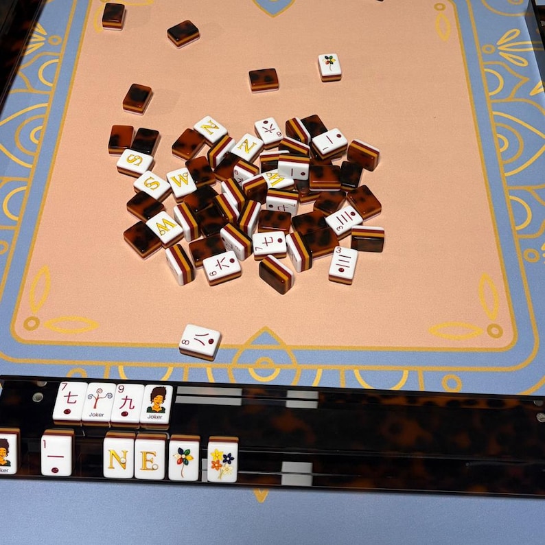 以下が含まれることがあります： ピンクと青のゲーム盤に散らばった、茶色と白の麻雀牌のクローズアップ。牌には様々な記号や文字が描かれています。セットには、追加の牌が入ったラックも含まれています。