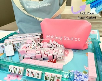 May include: A complete Mahjong set with pink and white tiles, dice, and racks. Includes a blue tote bag and a pink pouch, both with "Mahjong Studios" printed on them. The set is displayed on a floral patterned surface. Text on the image reads "Pick Your Rack Color!"