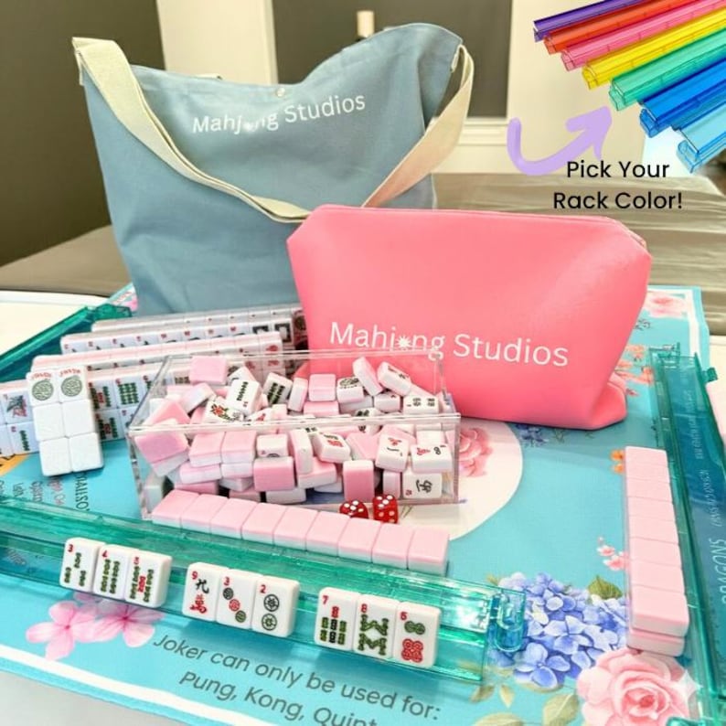 May include: A complete Mahjong set with pink and white tiles, dice, and racks. Includes a blue tote bag and a pink pouch, both with "Mahjong Studios" printed on them. The set is displayed on a floral patterned surface. Text on the image reads "Pick Your Rack Color!"