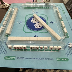 May include: A complete Mahjong set with white tiles and a light blue playing surface. The set includes dice and clear plastic tile racks. The text on the surface reads "Walls Left" and "Play Right".