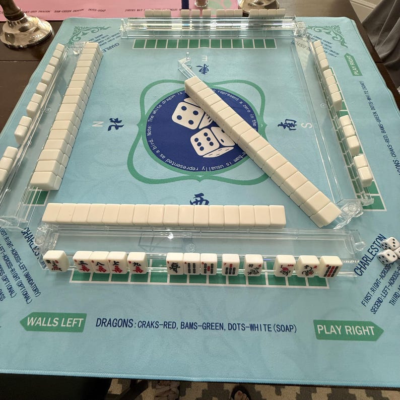 May include: A complete Mahjong set with white tiles and a light blue playing surface. The set includes dice and clear plastic tile racks. The text on the surface reads "Walls Left" and "Play Right".