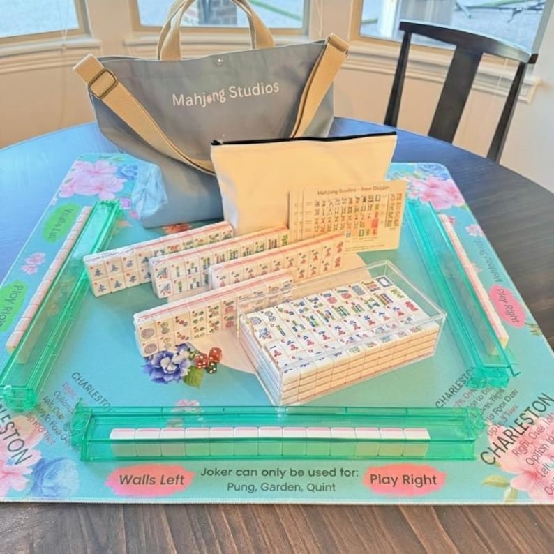 May include: A Mahjong set featuring a light blue tote bag with "Mahjong Studios" printed on it. The set includes tiles, racks, dice, and a playing surface with floral designs and the text "Charleston".