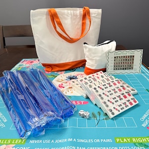 Puede incluir: Un juego completo de Mahjong con fichas blancas con marcas rojas y verdes, dados y un tapete de juego azul. Incluye un bolso de mano blanco con asas y ribetes naranjas, y una bolsa más pequeña a juego.