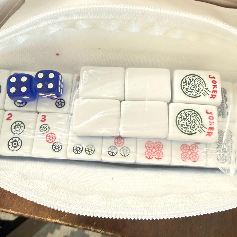 May include: A white zippered pouch containing a Mahjong set. The set includes white tiles with red and green markings, two blue dice with white dots, and tiles marked "Joker". The pouch is open, revealing the game pieces.