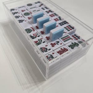 May include: A complete Mahjong set in a clear acrylic case. The white tiles feature red and green symbols and characters. Four light blue tiles are placed on top. The set is ready for play.