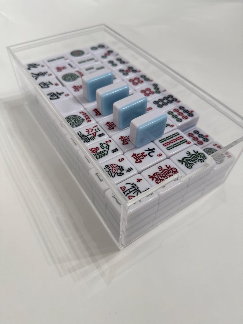 May include: A complete Mahjong set in a clear acrylic case. The white tiles feature red and green symbols and characters. Four light blue tiles are placed on top. The set is ready for play.