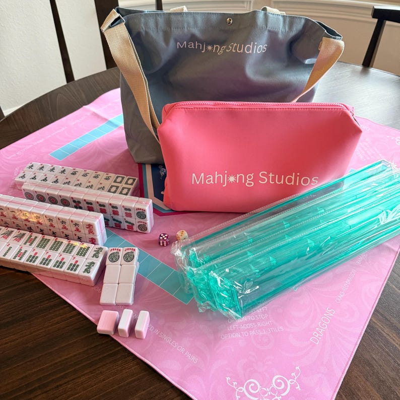 May include: A complete Mahjong set with tiles, dice, and accessories. Includes a pink zippered pouch and a gray tote bag, both with "Mahj*ng Studios" printed on them. The set is arranged on a pink mat with text.
