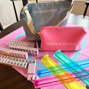 May include: A complete Mahjong set with a blue tote bag and a pink cosmetic bag, both with "Mahjong Studios" printed on them. The set includes tiles, dice, and colorful racks. The game is set on a pink and blue patterned mat.