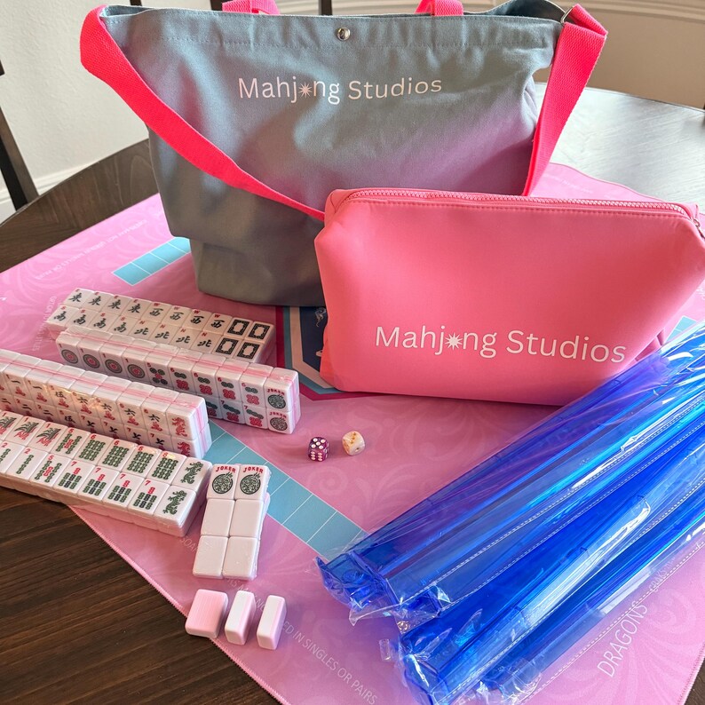 May include: A Mahjong set featuring a pink game board, tiles, dice, and accessories. Includes a grey tote bag and a pink zipped pouch, both with "Mahjong Studios" printed on them. Blue plastic sticks are also included.