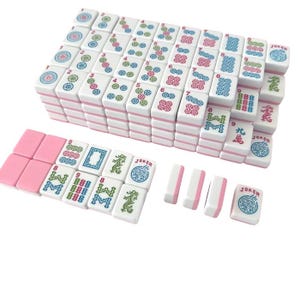 May include: A complete Mahjong set with white tiles featuring various symbols and numbers in red, green, and blue. The tiles have pink edges and are stacked neatly. Additional pink tiles and white tiles with pink edges are also included.