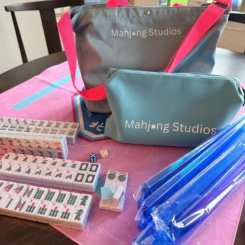 May include: A Mahjong set featuring tiles, dice, and accessories, alongside a grey tote bag and a matching cosmetic bag. Both bags have pink straps and the text "Mahjong Studios". The set is displayed on a pink and blue mat.