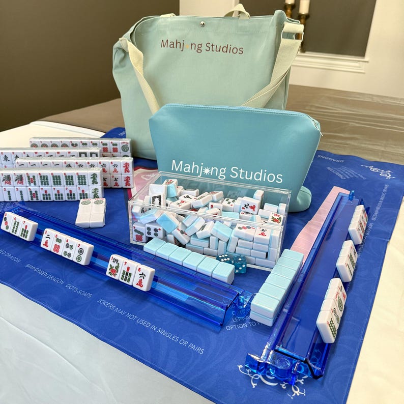 May include: A complete Mahjong set with blue and white tiles, dice, and racks, displayed on a blue cloth. The set includes a clear acrylic box and a matching blue zippered pouch and tote bag, all branded with "Mahj ng Studios".