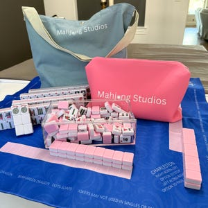 May include: A Mahjong set with pink and white tiles, a blue tote bag, and a pink cosmetic bag. The tote and cosmetic bag have the text "Mahjong Studios". The set includes a clear acrylic box of tiles and a blue playing mat.